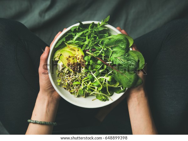 Green\
vegan breakfast meal in bowl with spinach, arugula, avocado, seeds\
and sprouts. Girl in leggins holding plate with hands visible, top\
view. Clean eating, dieting, vegan food\
concept