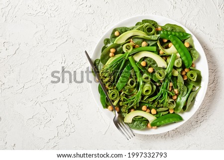 Green vegetable salad of sauteed tenderstem broccoli, snow peas, avocado, cucumber, chickpeas, baby spinach with olive oil dressing, served on a white plate on a white table, copy space, top view