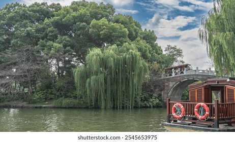 Green vegetation on the lake shore. Willow bent over the water. People are walking along the arched stone bridge. In the foreground is a fragment of a wooden boat with life buoys on the railing. - Powered by Shutterstock