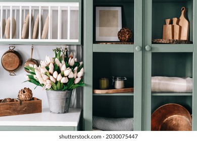 Green Vintage Cupboard With Glass Doors, Set Of Various Sized Wooden Cutting Boards And Tablecloths, Countertop With White Surface, Lots Of Flowers In Metal Bucket, Comfortable Kitchen Enviroment