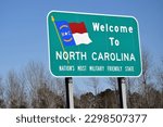 Green welcome to North Carolina road sign complete with the N.C. state flag on it. The background has blue sky and some trees without leaves as it was taken during a cold season. 