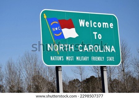 Green welcome to North Carolina road sign complete with the N.C. state flag on it. The background has blue sky and some trees without leaves as it was taken during a cold season. 