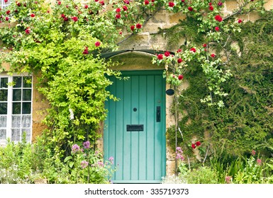 Green Wooden Doors In An Old Traditional English Stone Cottage Surrounded By Climbing Red Roses And Flowers