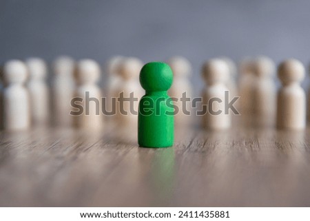 A green wooden peg doll stands out from a group of identical white peg dolls. Challenging conformity, standing out from the crowd and individuality concept.