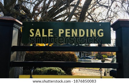 A green and yellow Sale Pending sign on a fancy post with trees, bushes, and a white picket fence in the background.