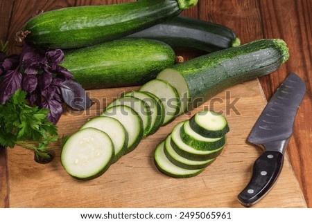 Green zucchini sliced into circles on the wooden cutting board against the same whole zucchini and different greens on the rustic table 
