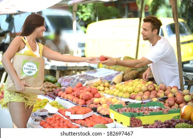Greengrocer owner of a small business at an open street market, handing out a fruit to a consumer, carrying a shopping bag with 100% organic certified label.