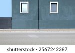 Greenish gray plaster wall with gutter in the middle, two windows by sides and black metal fence on left. Sidewalk and street in front. Background for copy space.