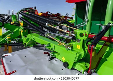 Green-painted farm implement viewed from a low angle showing hydraulic hoses, metal linkages, bolts, and painted steel panels under daylight. The scene