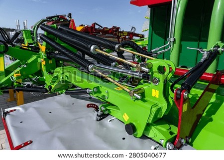 Green-painted farm implement viewed from a low angle showing hydraulic hoses, metal linkages, bolts, and painted steel panels under daylight. The scene