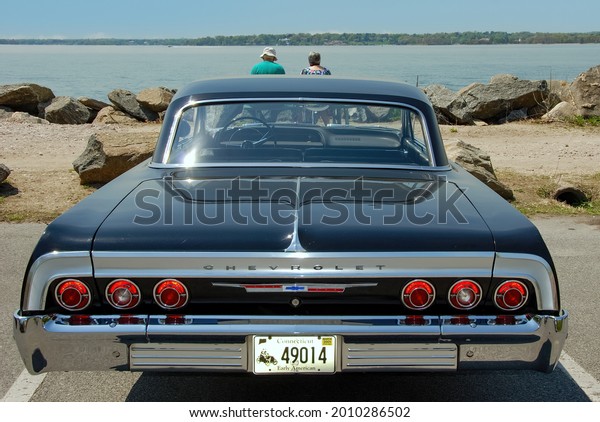 Greenwich, CT, USA-May\
2011; View of a classic Chevrolet Impala seen from the rear with\
two people in front of car on a bench overlooking the Long Island\
Sound on sunny day