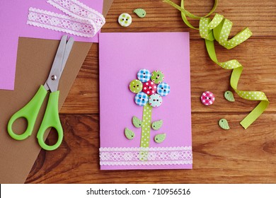 Greeting card with a flower made of wooden buttons, decorated with lace. Beautiful paper card crafts for mom's birthday or mother's day. Tools and materials on a table. Children's workplace. Top view - Powered by Shutterstock