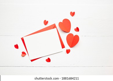 Greeting card mockup, white blank card with paper envelope and red parer hearts on wooden table background. Valentine's day greeting card, flat lay, top view