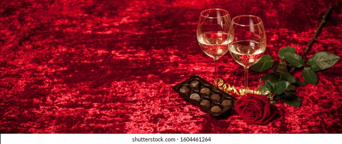 Greeting Card For Valentine's Day - Two Glasses With White Wine, A Rose And Chocolates On A Romantic Red Background. Concept - Valentines Day, Holiday, Mothers Day, Banner.