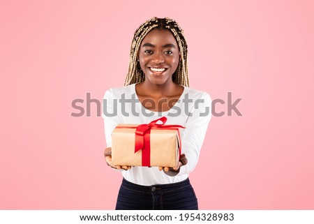 Greeting And Celebration. Portrait of positive cheerful African American lady holding wrapped gift box, giving present to the camera, standing isolated over pink color studio background, free space