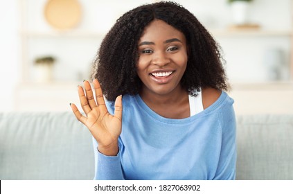 Greeting Concept. Portrait Of Smiling Beautiful African American Woman Waving Hello With Hand Looking At Camera, Sitting On Couch At Home. Black Lady Saying Hello And Laughing, Blurred Background