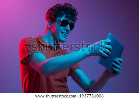 Greeting friend online. Young man in sunglasses chating isolated on multicolored background in neon light. Concept of human emotions, facial expression, youth culture, digital life. Copy space for ad.