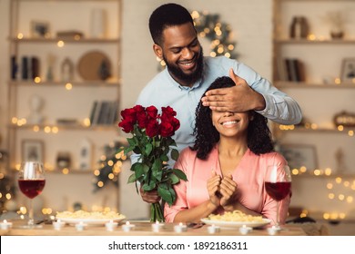 Greeting With Holidays. Smiling Black Man Covering His Woman Eyes And Giving Her Bunch Of Red Roses, Making Surprise To Beautiful Lady. African American Couple Celebrating Together At Home Or Cafe