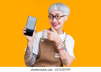 greeting old senior asian female business owner in apron hand gesture show smartphone blank screen with joyful presentation,asia woman tattoo wear glasses barista hand pointing  show cellphone online