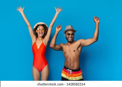 Greeting summer time. Happy african american millennial couple in swimwear jumping on blue studio background