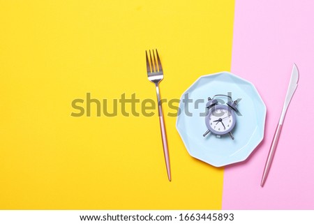 Grey alarm clock,fork and knife in empty blue plate on yellow and pink background.Concept of intermittent fasting, lunchtime, diet and weight loss.Top view,flat lay,minimalism.