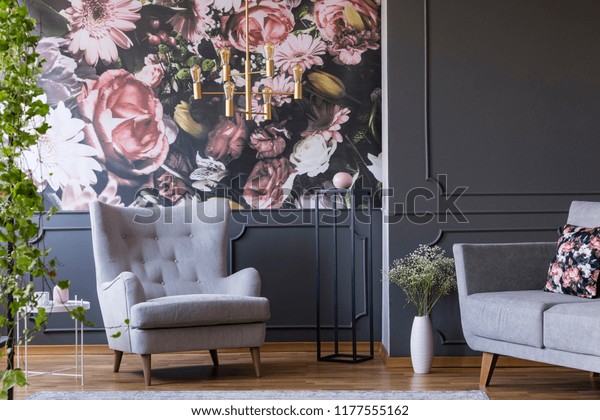 Grey armchair against flowers wallpaper in dark living room interior with sofa and plant. Real photo