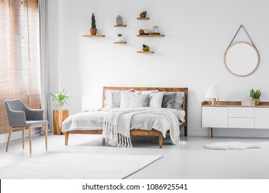 Grey Armchair Near Bed With Knit Blanket In White Bedroom Interior With Cupboard And Plants