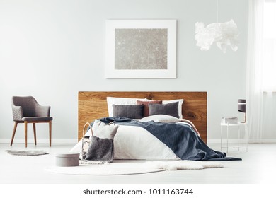 Grey Armchair Standing Next To Wooden Bedhead Bed With Pillows In Bright Bedroom Interior With Silver Poster On The Wall