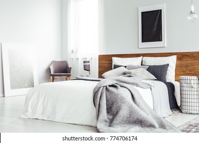 Grey blanket on bed with wooden bedhead in simple bedroom interior with dark poster and chair under window - Shutterstock ID 774706174
