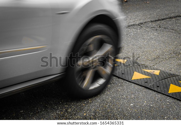 Grey car Driving Up to and Just\
Connecting with Yellow and Black Striped Speed Bump in\
Ourense.