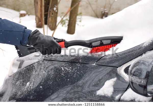 Grey\
car outside covered with snow and ice. man in blue jacket and\
leather black gloves shoveling snow from window glass and\
windscreen by red plastic brush. Winter season\
work.