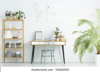 Grey Chair At Desk Against White Wall With Poster In Home Office With Plants On Wooden Shelves