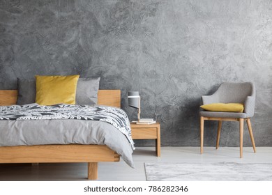 Grey chair and lamp on wooden nightstand in dark bedroom with concrete wall and yellow pillow on bed
