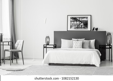 Grey chair at table near white bed in spacious contrast bedroom interior with black and white poster