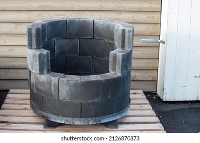 Grey circular porous backyard pavers stacked to make a homemade fire pit. The bricks are fireproof resistance stones with an opening in the front. There's a wooden tan wall in the background. 