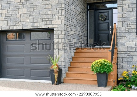 A grey colored modern garage door, two large flower pots with mums and cascading flowers at the end of brown wooden stairs to a black panel door. The exterior wall of the house is grey colored brick. 