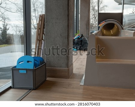 A grey crate holds blue floatation boards beside a large window in a contemporary water park facility, emphasizing clean organization for leisure activities and swimming pool equipment
