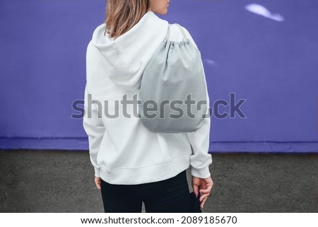 Grey drawstring pack template, mockup of bag for sport shoes on woman's shoulder standing on a violet or very peri background.