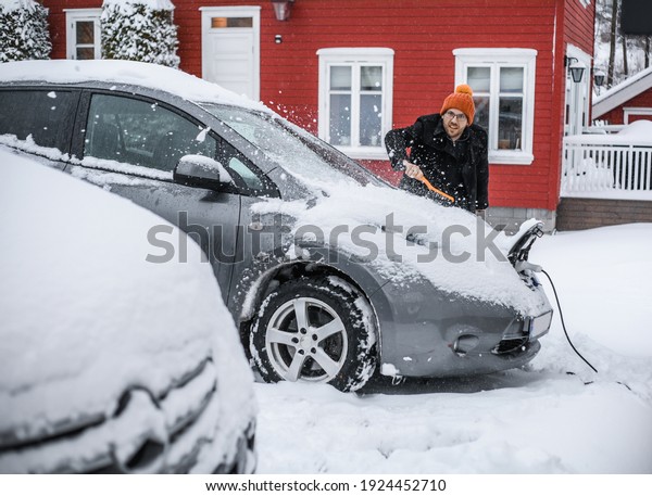 grey   electric car with
Scandinavian house in the background. A nerdy guy with brush is
removing snow.