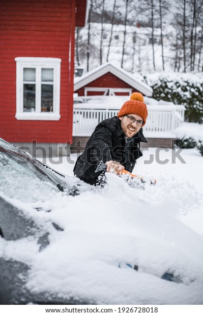 grey   electric car with\
Scandinavian house in the background. A nerdy guy with brush is\
removing snow.