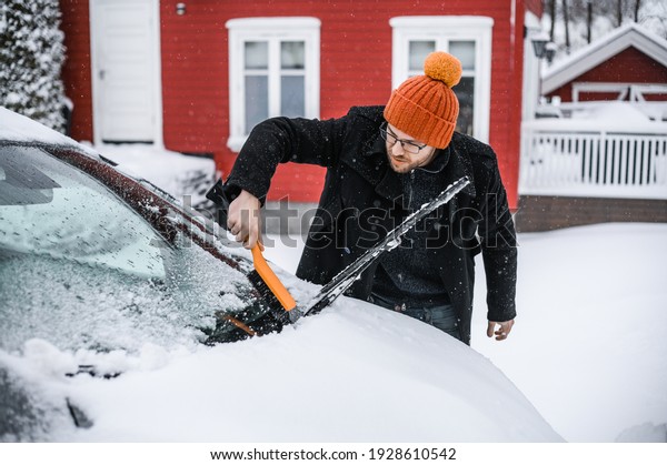 grey   electric car with\
Scandinavian house in the background. A nerdy guy with brush is\
removing snow.