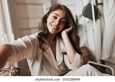 Grey eyed woman in white blouse taking selfie. Charming girl with wavy hair widely smiles and makes photo while sitting on wooden floor - Shutterstock ID 1925288984