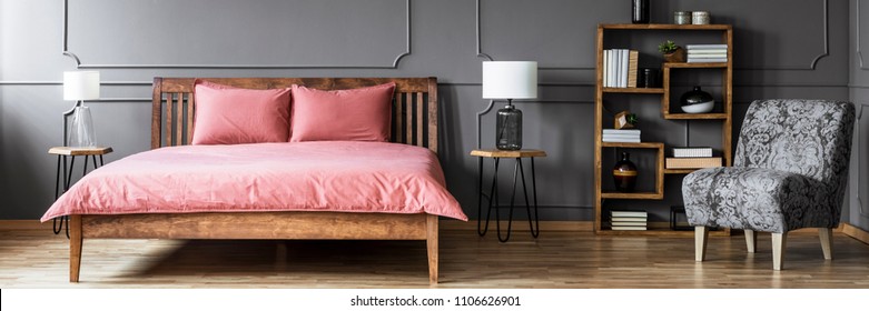 Grey floral armchair and wooden rack with books and decor standing in dark bedroom interior with pink sheets on bed placed between two hairpin tables with lamps