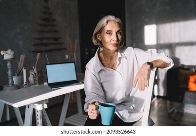 Grey Hair Caucasian Businesswoman With Cup Of Tea Or Coffee Sitting On Chair And Looking At Camera. Small Business And Entrepreneurship. Modern Successful Woman. Dark Art Studio With Sunny Lighting