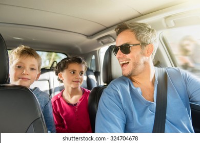 A Grey Hair Father With Beard And Sunglasses Is Laughing With His Two Kids. They Are In The Car Leaving For The Weekend. The Mother Is In The Background, Looking At Her Phone.