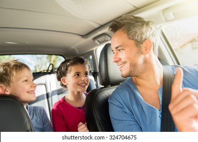 A Grey Hair Father With Beard Is Talking With His Two Kids In The Car. The Ten Years Old Brother And Sister Are Sitting In The Back, They Are Wearing Long Sleeves Shirts. The Father Have His Seat Belt