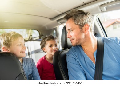 A Grey Hair Father With Beard Is Talking With His Two Kids In The Car. The Ten Years Old Brother And Sister Are Sitting In The Back, They Are Wearing Long Sleeves Shirts. The Father Have His Seat Belt