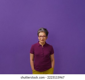 Grey Hair Senior Woman Posing With Hands In Pockets Looking At Camera Wearing Glasses, Purple Polo And Mustard Jeans, Isolated On Dark Velvet Background. Healthcare, Aged Beauty Concept. Copy Space. 