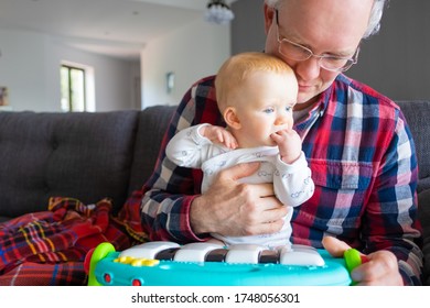 Grey Haired Grandpa Holding Baby In Arms At Home, Teaching Little Granddaughter To Play Toy Piano. Family Or Learning Games Concept