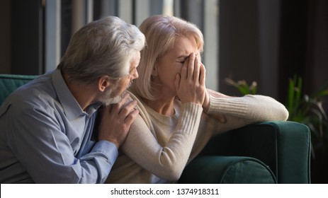 Grey haired man supporting crying unhappy mature woman, health problem, disease, bad news, loving middle aged husband hugging old wife by shoulders, sitting on couch at home, horizontal banner